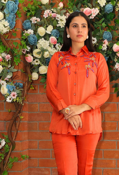 the Shimmer Glimpse coord set from Destiny by Anjali. This vibrant designer western wear comes in a striking bright orange, made from premium bombarg satin fabric. The shirt features intricate zardozi hand embroidery work, adding a touch of regal elegance. Fully lined for comfort, this set is perfect for all-weather and occasion wear, blending traditional craftsmanship with a modern aesthetic. Stay ahead of the trending fashion curve with this sophisticated and versatile floral coord set